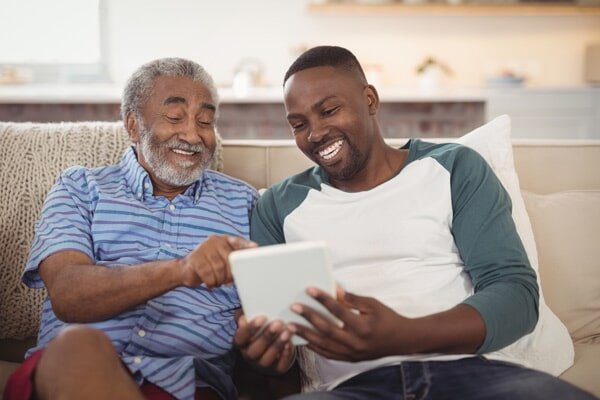 older man and young man sitting on a couch smiling and looking at a white tablet